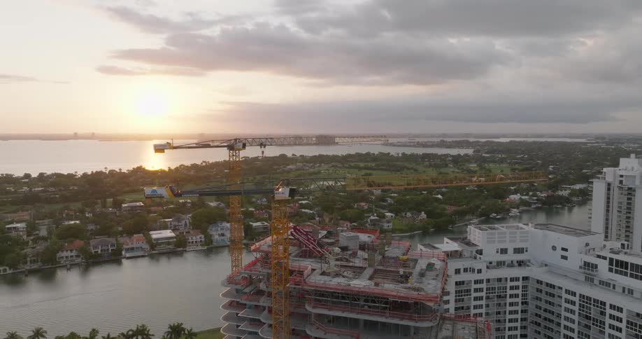 An aerial orbit reveals a high-rise building under construction with cranes overlooking the Miami bay and skyline at sunrise