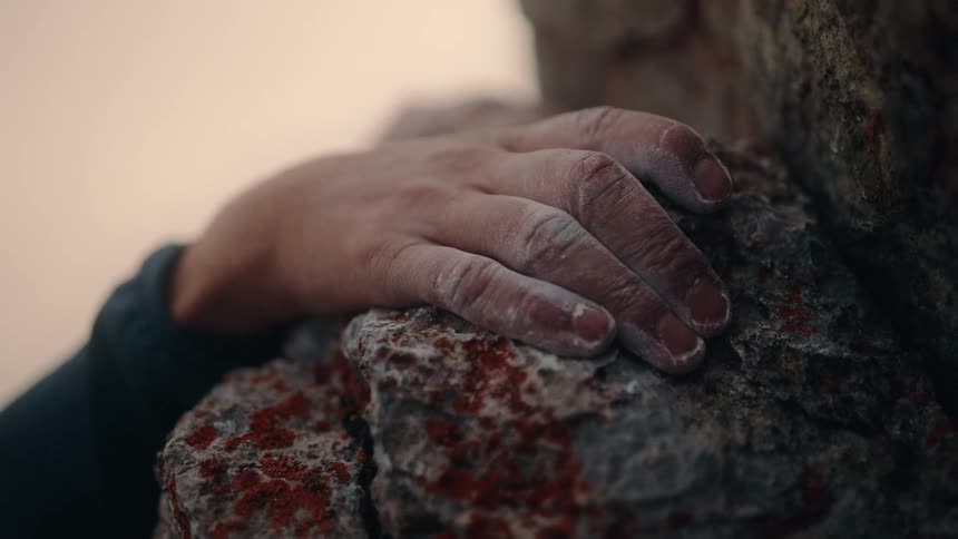 A climber's hand, covered in chalk, slowly and firmly grips a lichen-covered rock hold in a dramatic close-up
