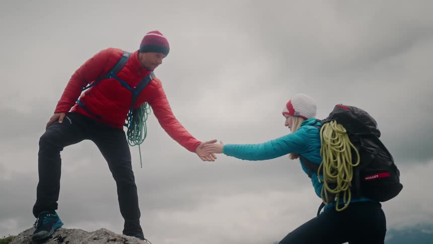 A male hiker offers a helping hand to his female partner as they climb a rocky mountain together in slow motion
