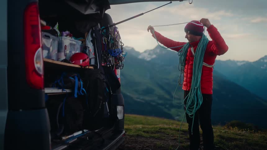 A male climber coils his rope next to his adventure van while preparing for a climb in the mountains at sunrise