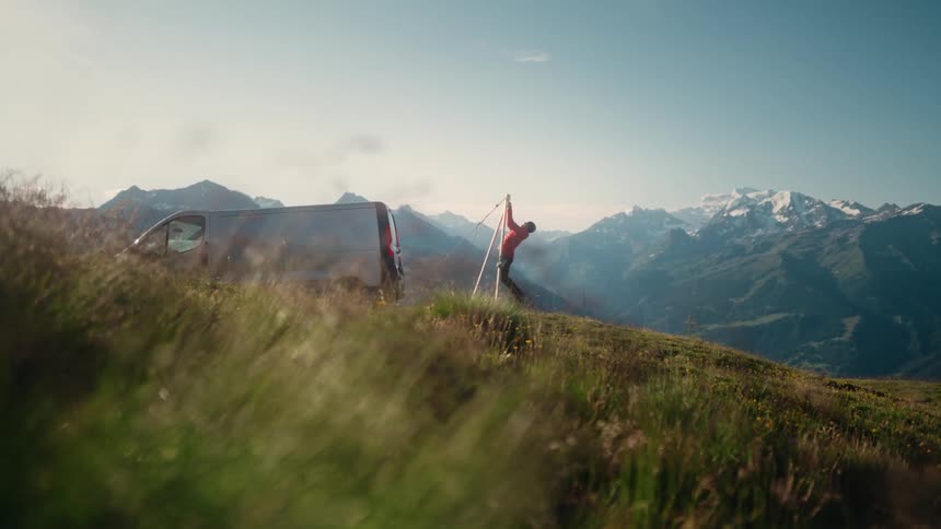 A person on stilts practices balancing on a grassy mountain top next to their camper van with a stunning view of snow-covered peaks