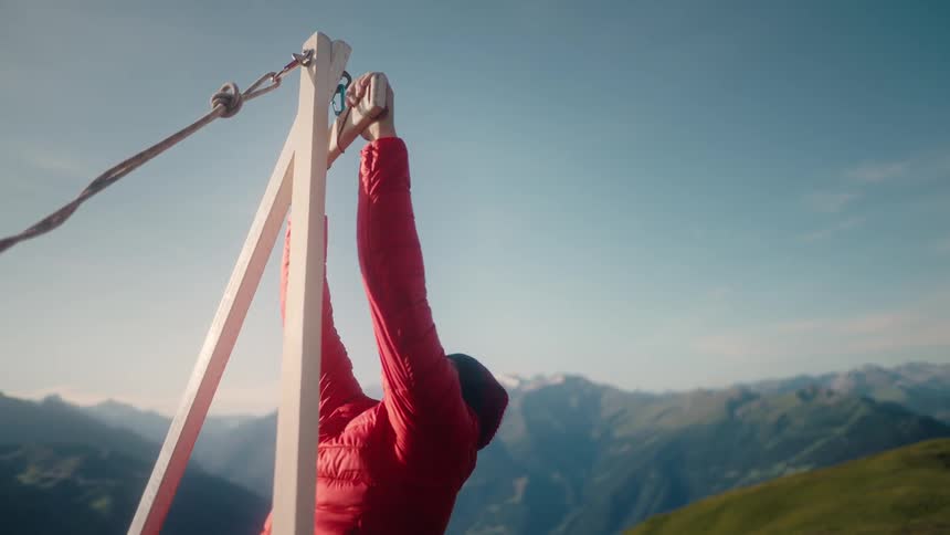 A man in a red jacket sets up and tightens a slackline anchor system on a mountaintop with a beautiful mountain range view in the background