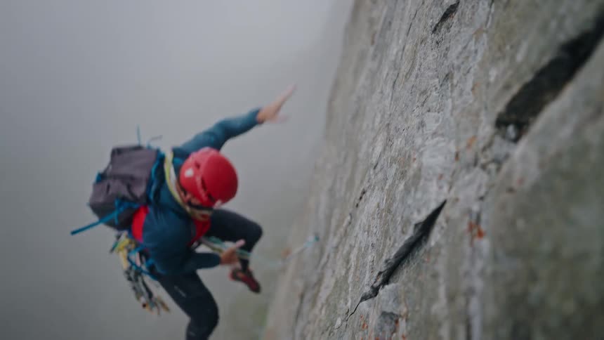 A mountaineer in a red helmet takes a controlled fall in slow motion while rock climbing a steep cliff face in the fog