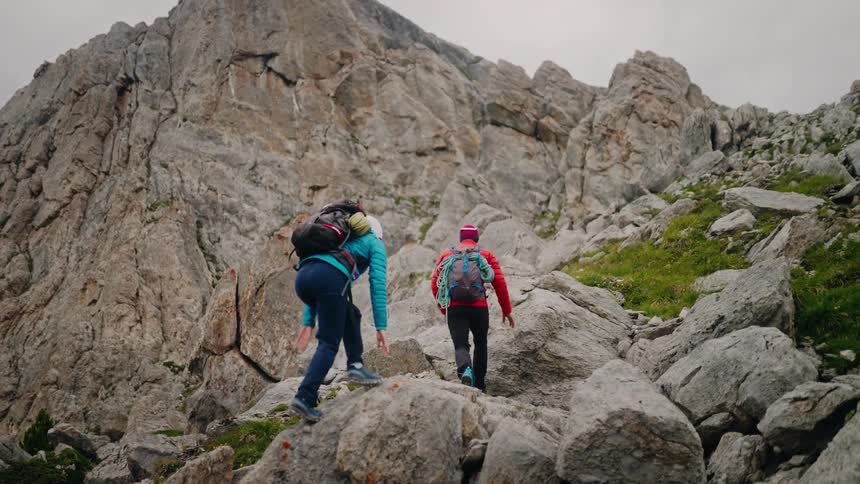 Two female hikers with full backpacks and climbing gear carefully scramble up a steep and rocky mountain trail