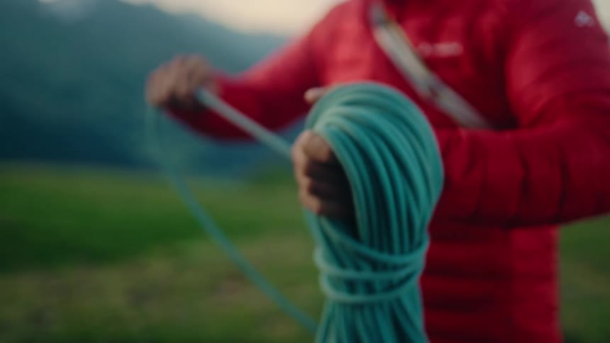 A mountain climber wearing a red jacket carefully coils a green climbing rope in slow motion in preparation for an ascent