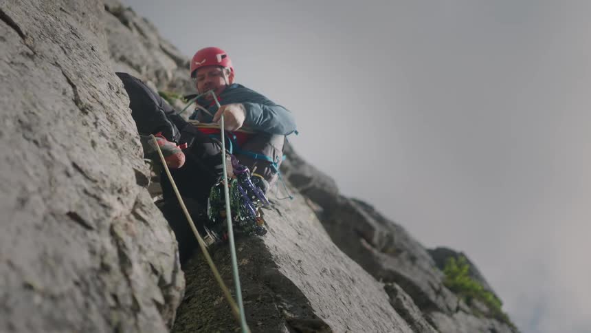 An experienced male rock climber pulls up his rope while sitting on a narrow mountain ledge
