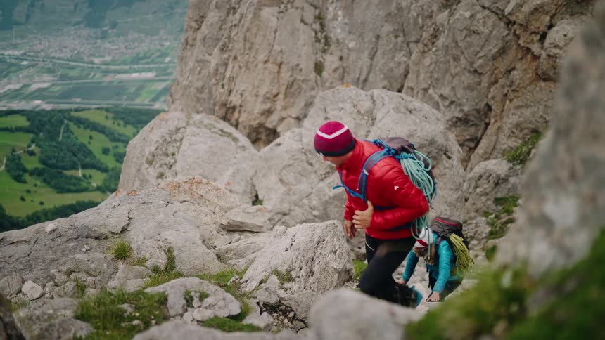 A male and female mountaineer team with full climbing gear carefully ascend a steep, rocky alpine trail