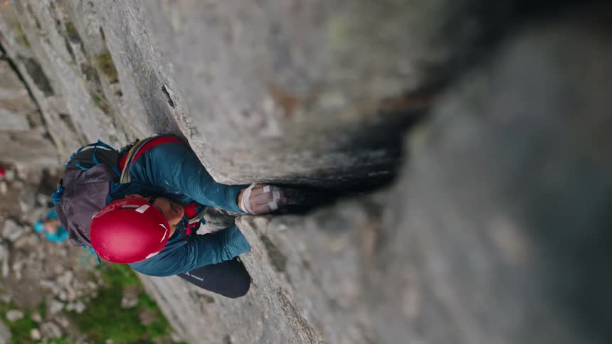 A determined climber in a red helmet carefully navigates a crack while scaling a steep rock face in this overhead slow-motion shot
