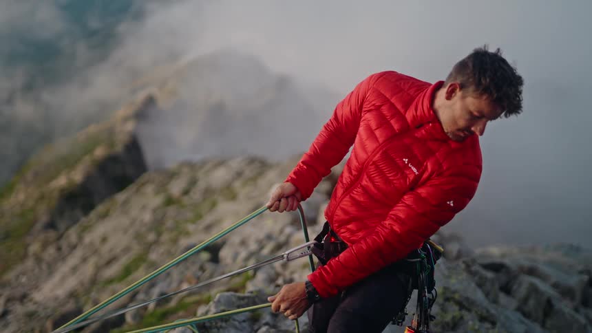 A male rock climber in a red jacket belays from a foggy mountain summit during an alpine ascent