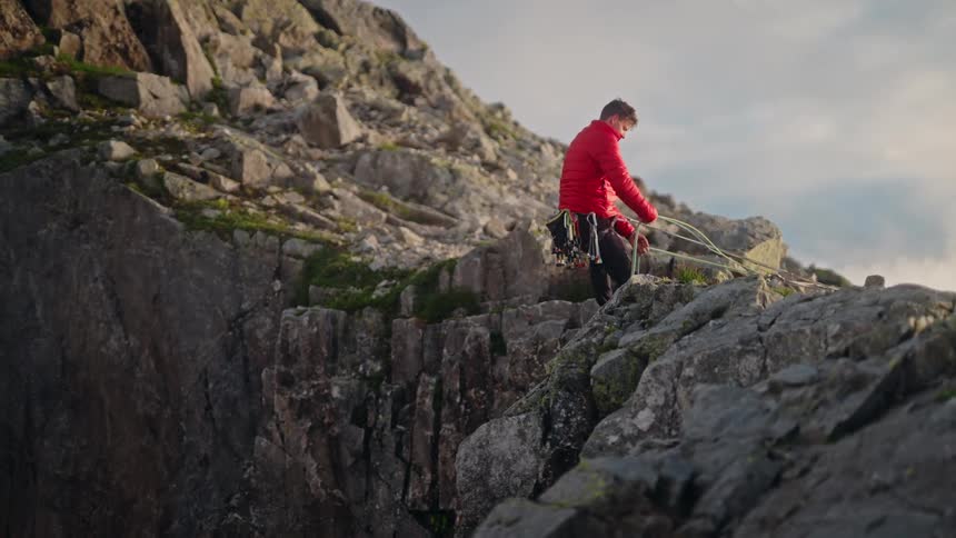 A mountain climber in a red jacket pulls in his safety rope while standing on a rugged, rocky ridge