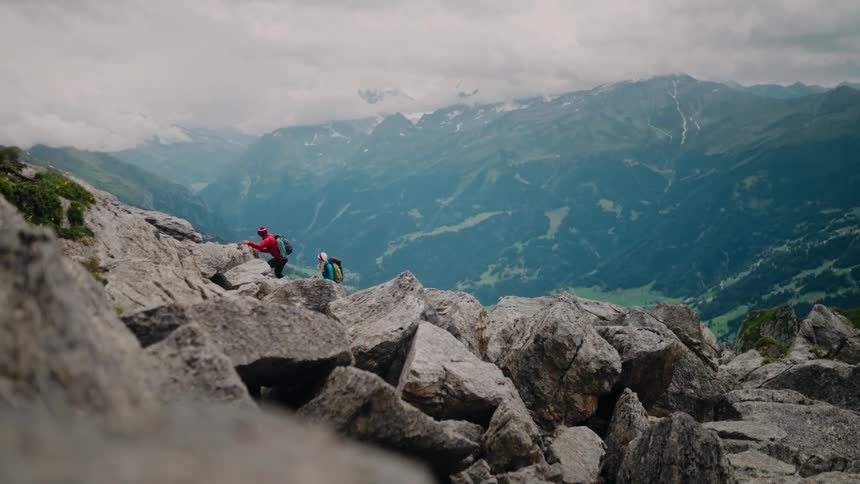 A man gives a helping hand to a woman as they carefully climb a rocky mountain trail with a vast, cloudy alpine valley below them
