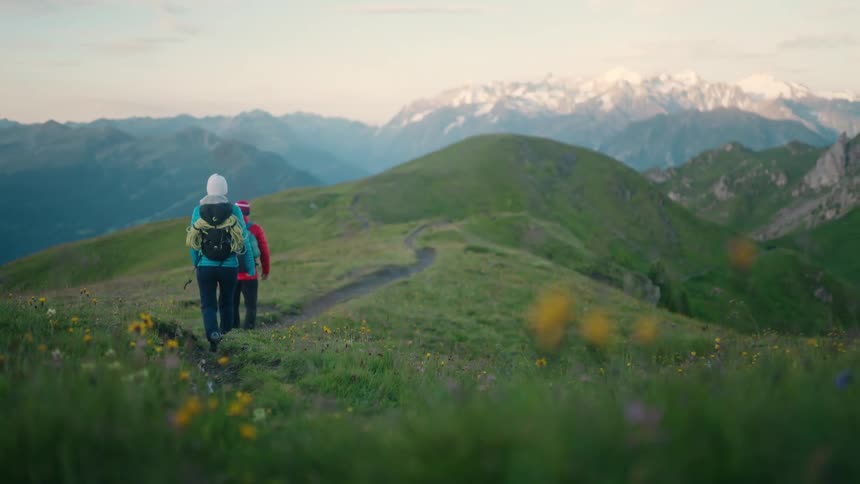 A couple hikes along a scenic trail on a lush green mountain ridge, with majestic snow-capped peaks visible in the distance at dawn