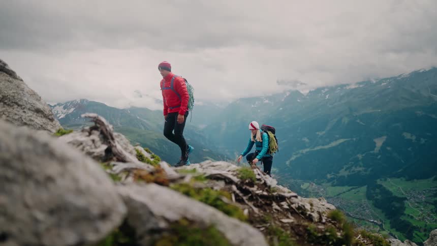 A man and a woman with backpacks and climbing gear carefully hike up a steep, rocky mountain ridge overlooking a beautiful green valley below