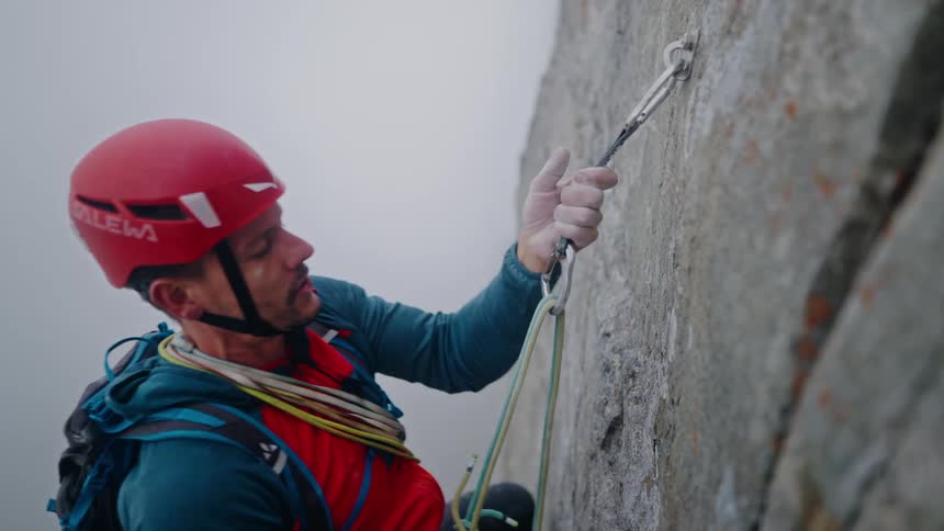 A focused rock climber wearing a helmet and backpack attaches his safety rope to a carabiner while scaling a steep, foggy mountain face