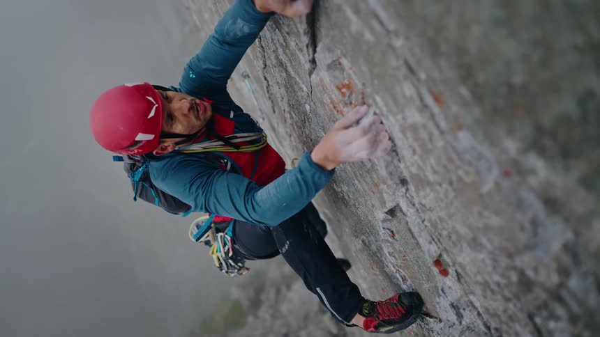 Vertical high-angle slow-motion shot of a determined man in a red helmet carefully rock climbing up a sheer cliff face