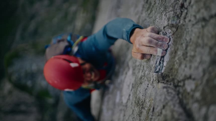 A determined man with a red helmet climbs a steep rock face, smiling as he finds a good grip in slow motion