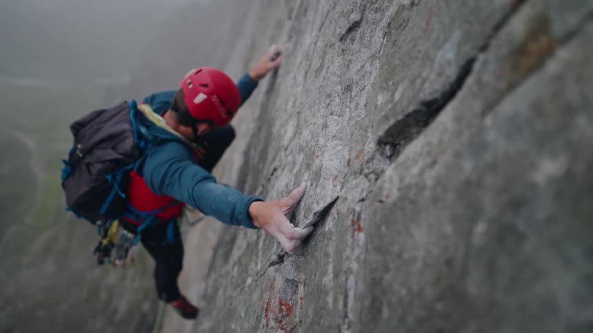 An athletic man in a helmet slowly and carefully ascends a sheer rock face, then rests his head against the mountain in the fog