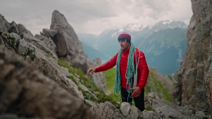 A climber in a red jacket prepares his climbing rope on a rocky mountain ridge, with snow-capped peaks visible in the distance