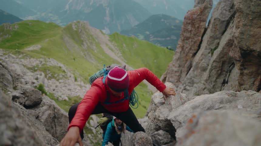 A man and woman with backpacks and climbing gear carefully scale a steep, rocky mountain path in slow motion