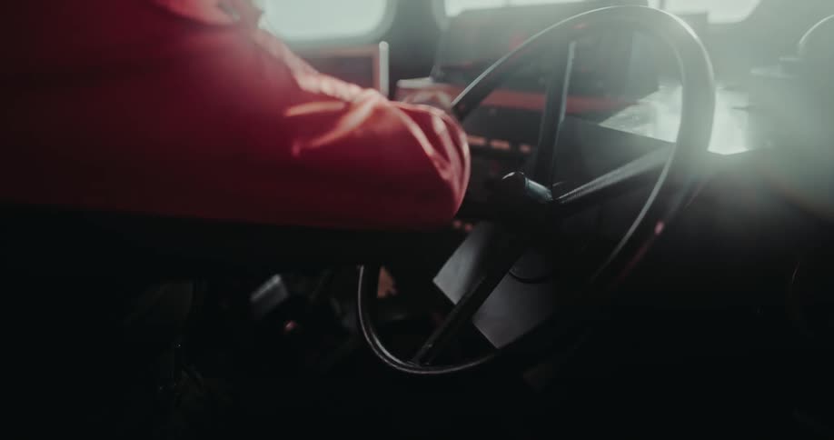 A boat captain in a red jacket steers the vessel from the helm inside a sun-drenched wheelhouse