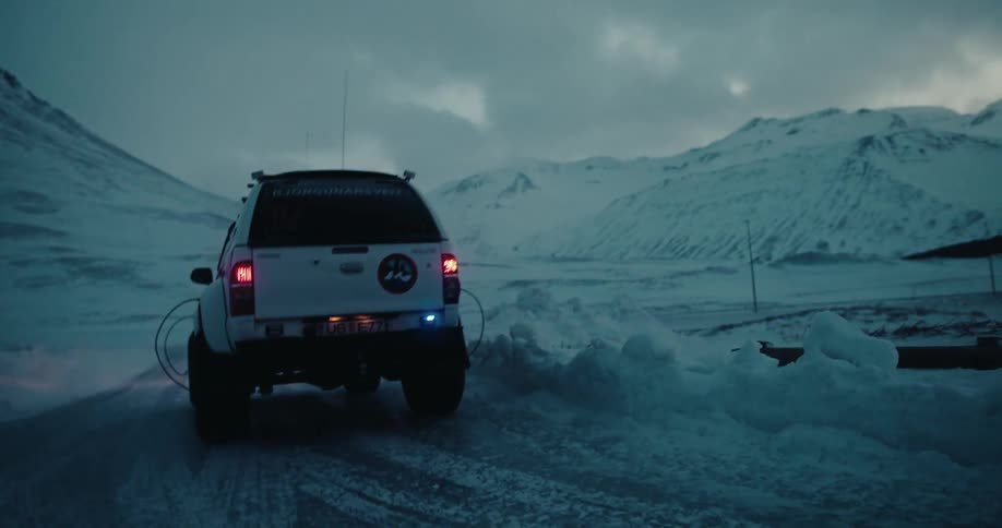 A search and rescue truck with flashing lights drives away into the remote, snow-covered mountains of Iceland