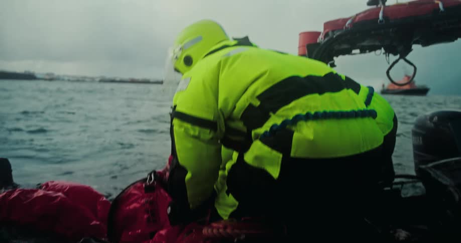 A search and rescue team in full gear performs a water rescue, pulling a person in a red survival suit from the ocean in slow motion