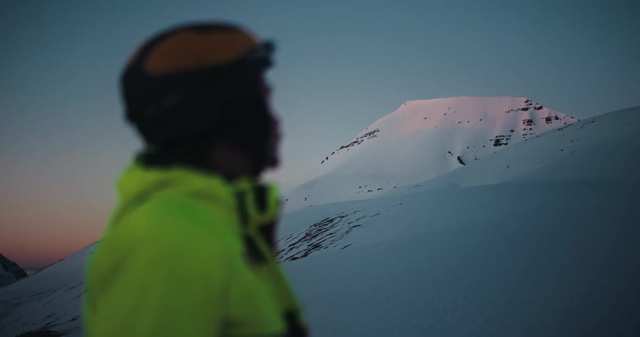 A person in a helmet and bright jacket watches the sunset over a vast, snowy mountain landscape in a slow focus reveal
