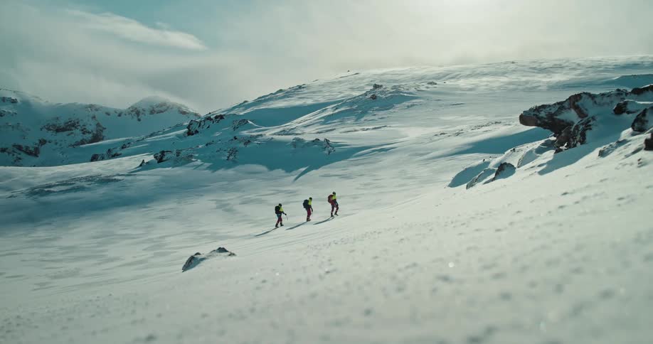 A team of three mountaineers with backpacks slowly ascends a vast, sun-drenched snowy mountain landscape