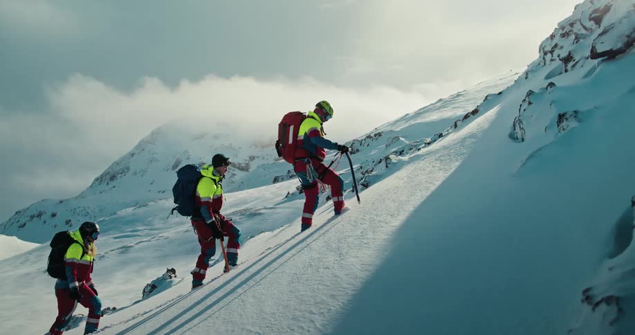 A mountain rescue team of three climbers in full gear carefully ascends a steep, snowy slope using ice axes in slow motion