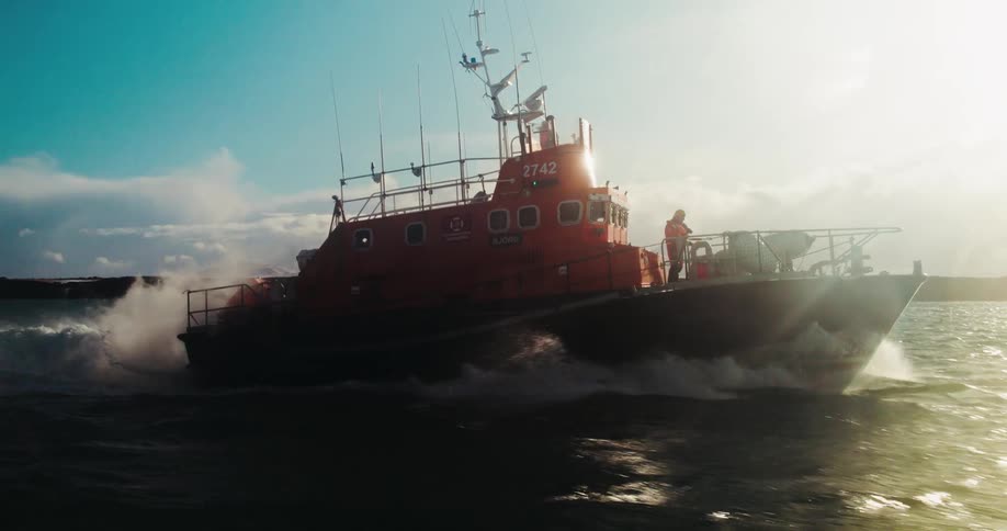 A bright orange search and rescue lifeboat speeds across the open ocean in dramatic slow motion as the sun shines brightly