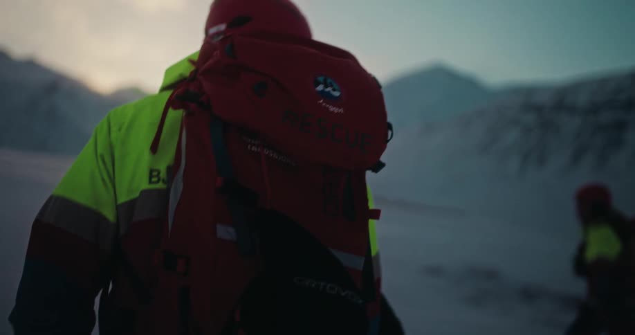 A mountain search and rescue worker hikes through a snowy, mountainous landscape at dusk