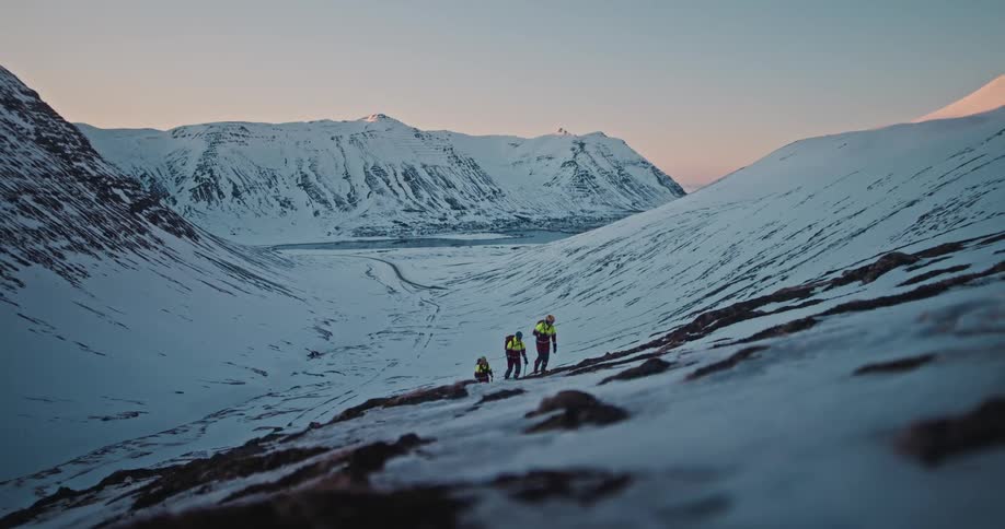 Three adventurous hikers with backpacks and poles ascend a steep snowy mountain during a beautiful sunrise