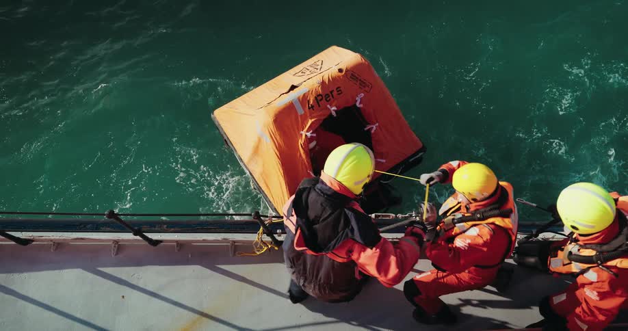 A top-down slow-motion view shows a ship's crew in safety gear deploying an inflatable life raft during a man overboard drill at sea