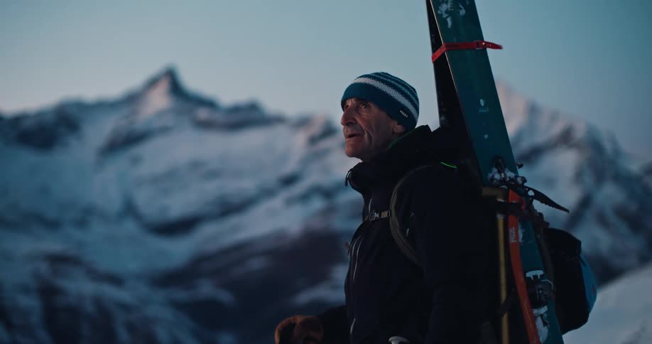 A seasoned backcountry skier with skis on his back pauses to admire the vast, snowy mountain range at dawn