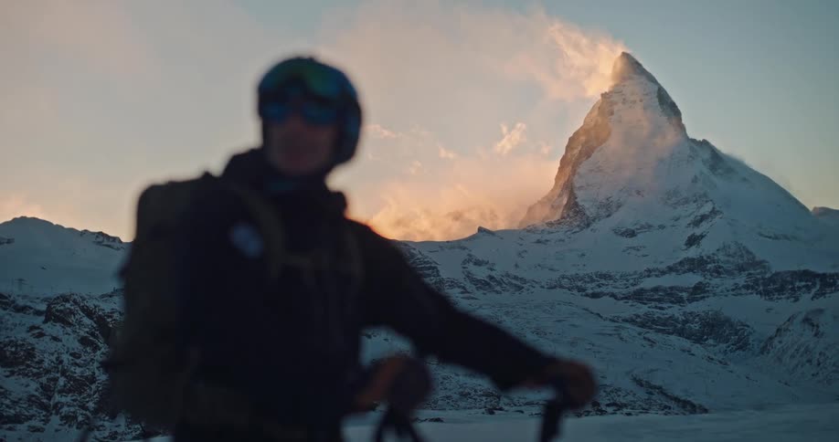 A silhouetted skier with a backpack watches the majestic Matterhorn peak as golden light from the setting sun illuminates the swirling clouds