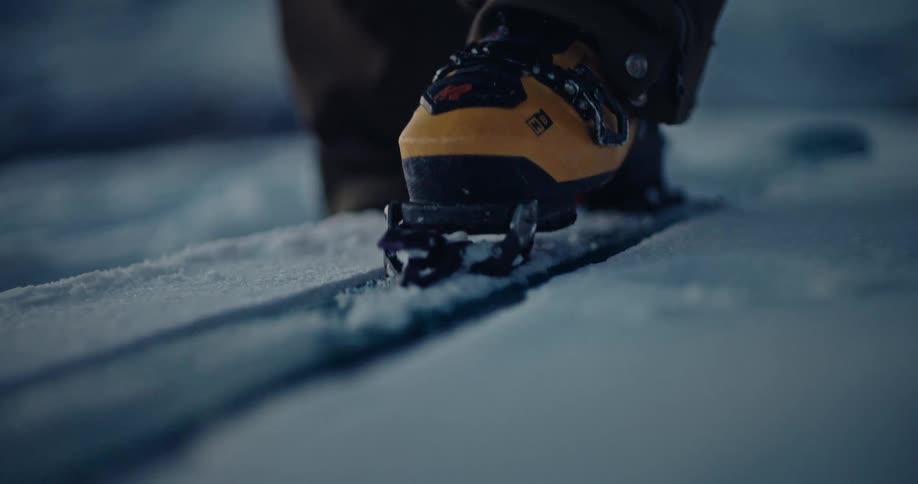 A backcountry adventurer secures their boot into a splitboard binding before hiking through deep, fresh snow in slow motion