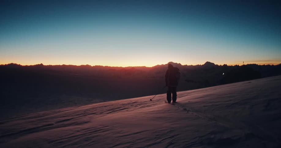 A solo backcountry skier treks up a windy, snowy mountain ridge, silhouetted against the rising sun