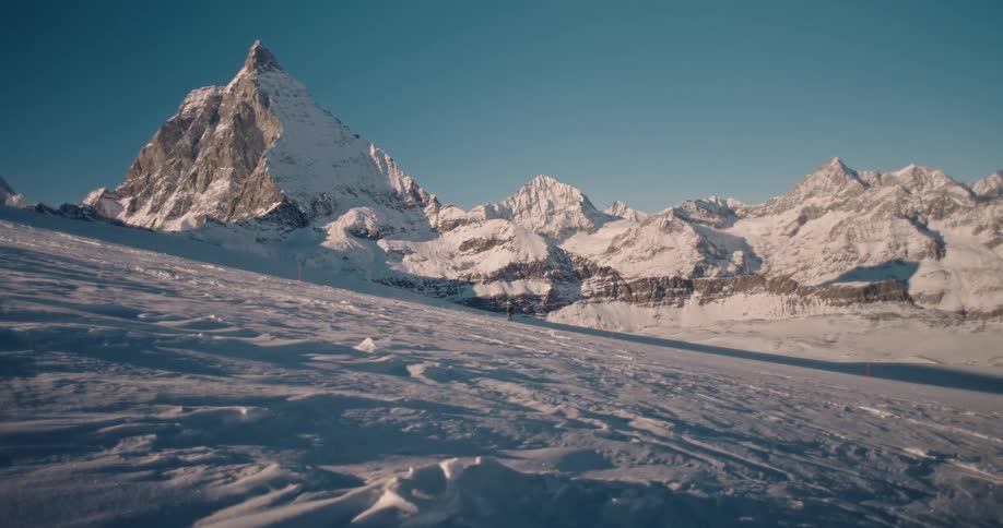 A lone skier gracefully descends a vast snowy slope, with a breathtaking panoramic view of the Matterhorn and the surrounding Alps under a clear blue sky