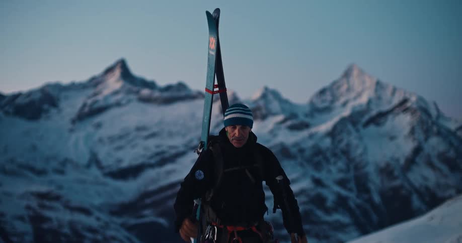 A lone mountaineer with skis on his back ascends a snowy peak and pauses to admire the vast mountain range at sunrise