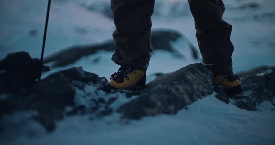 A close-up slow motion shot of a mountaineer's boots carefully stepping on snowy rocks at dusk