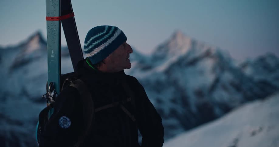 A senior backcountry skier with skis on his back pauses to admire the beautiful snow-covered mountain landscape at dusk