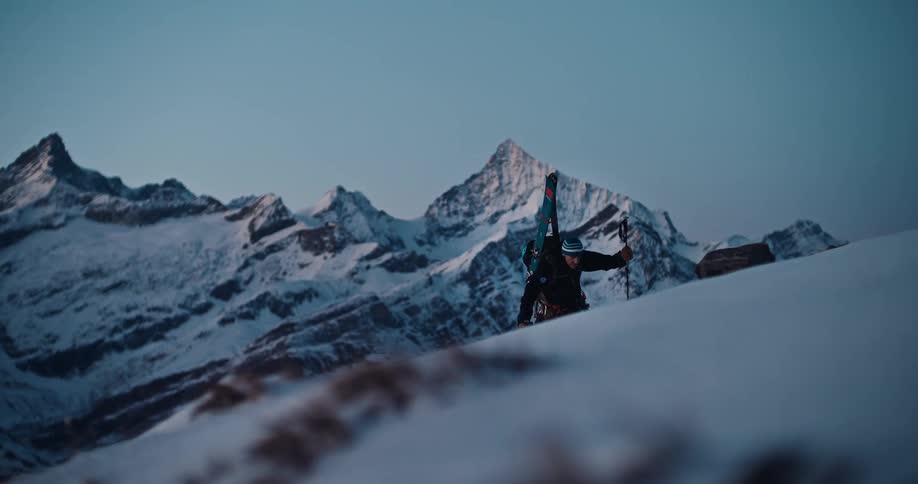 A male ski mountaineer carrying his skis hikes up a steep snowy ridge at sunrise with a stunning mountain range in the background
