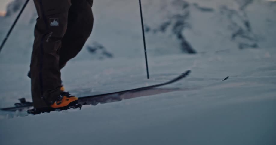 A backcountry skier ascends a snowy mountain, captured in cinematic slow motion as the sun sets behind the peaks