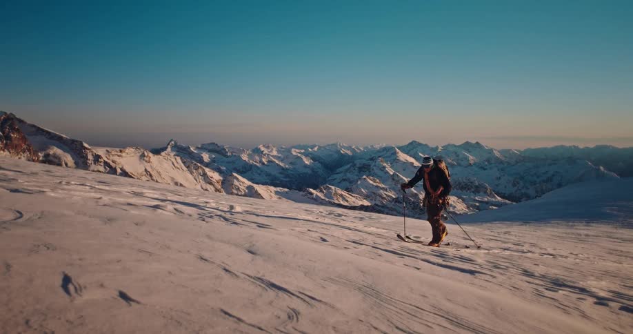 A lone ski mountaineer treks up a vast, snow-covered mountain slope at sunrise with a stunning mountain range in the background