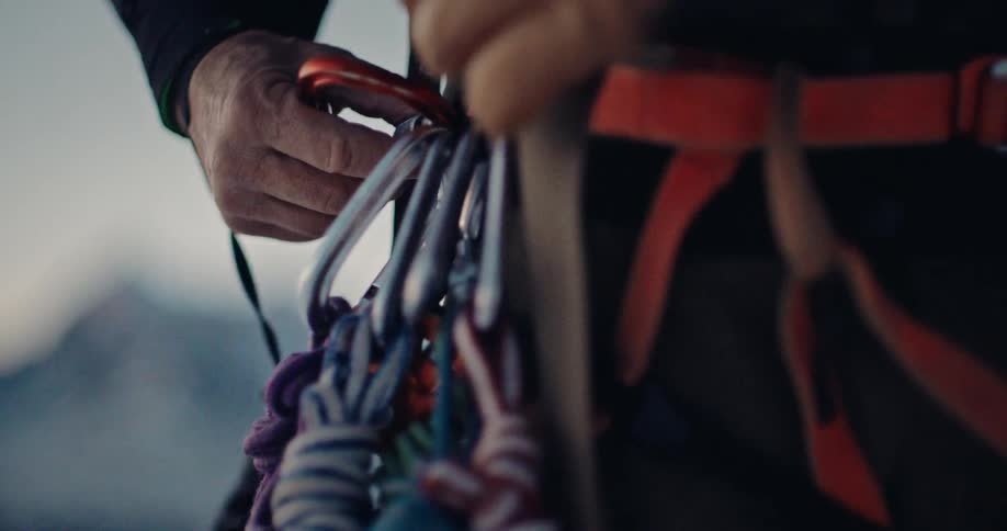 An alpinist meticulously prepares their climbing gear, securing a carabiner and rope to their harness in slow motion