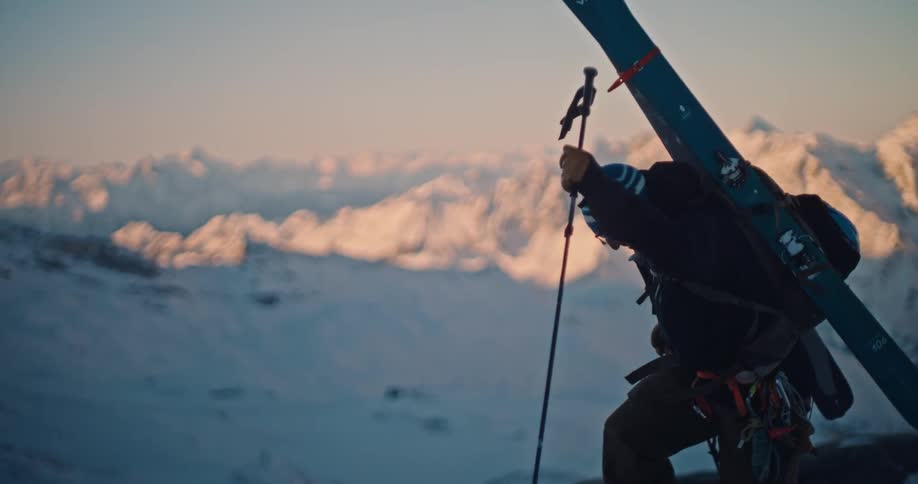 Slow motion of a determined mountaineer carrying skis while hiking up a steep, snowy mountain slope against a backdrop of breathtaking sunlit peaks