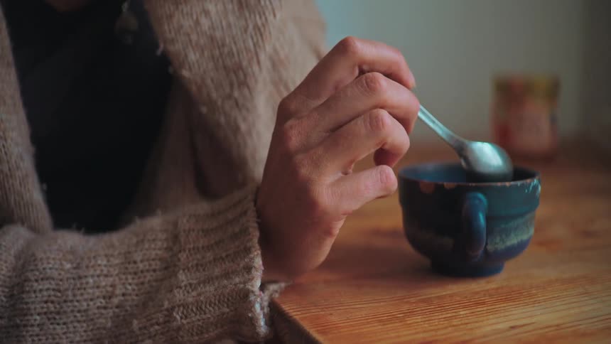A person wearing a cozy knit sweater slowly stirs a steaming hot beverage in a rustic ceramic mug on a wooden table