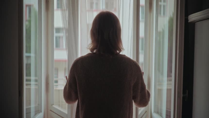 A silhouetted person in a sweater slowly opens the sheer curtains on a bright window, looking out into the city apartment courtyard
