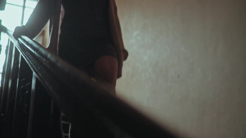 A mysterious woman in a cardigan walks up a dark, moody staircase, her hand gliding along the banister