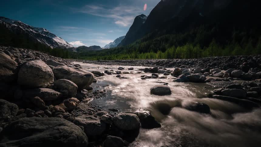 A beautiful long-exposure timelapse of a river winding through a rocky valley surrounded by snow-capped mountains and green forests